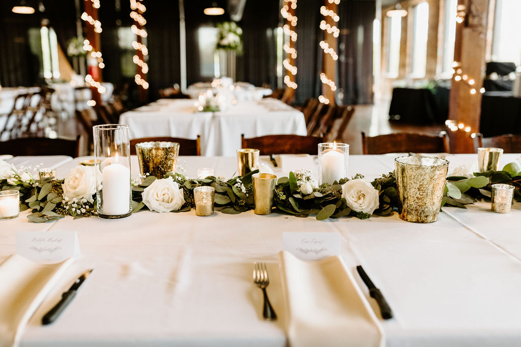 reception table runner consisting of greenery and white floral with gold accents and candles.