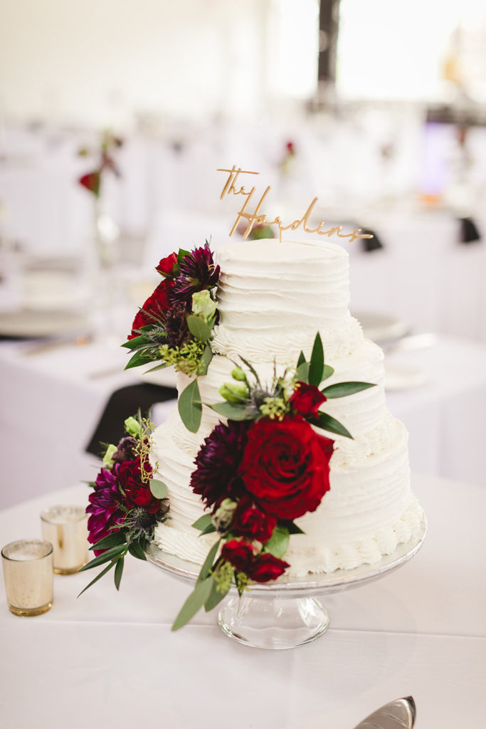 The wedding cake was three tiers of white layers accented with red and dark purple flowers and greenery with a gold cake topper that said "The Hardins" 