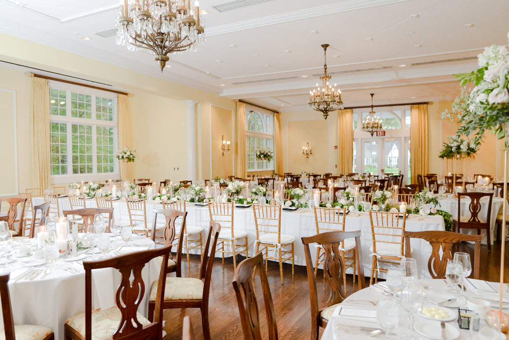 The wedding reception decorations inside the woodstock country club. The decor included white table linen, wood and gold chairs, flower centerpieces that ran along the center of the center kings table and tall flower centerpieces on round tables. 