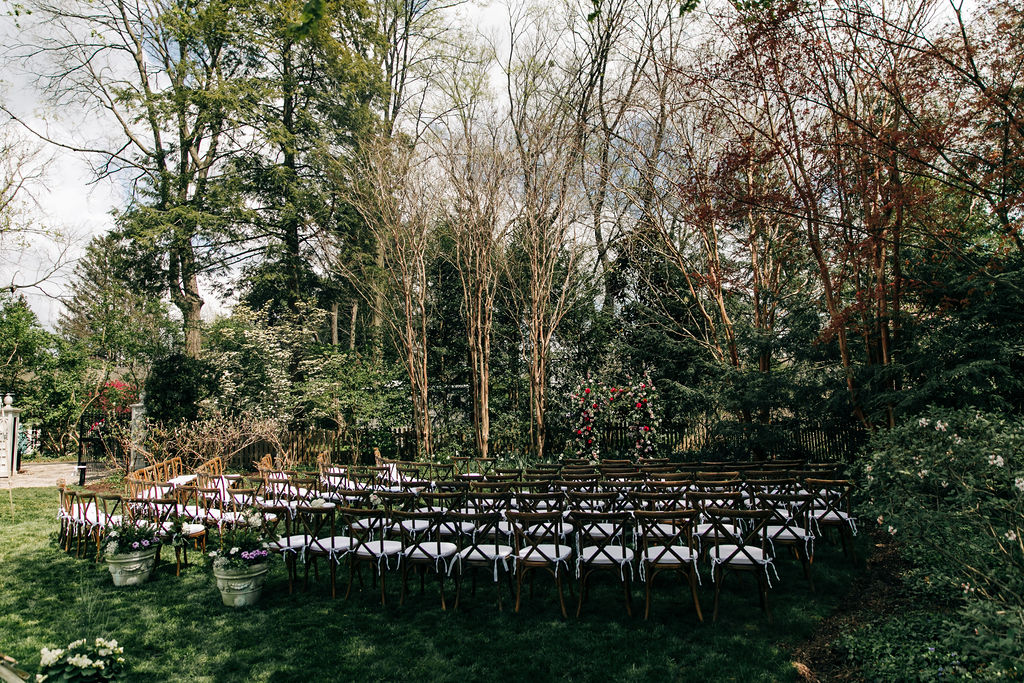 backyard wedding ceremony with wood cross back chairs, white cushions, and big flower arch