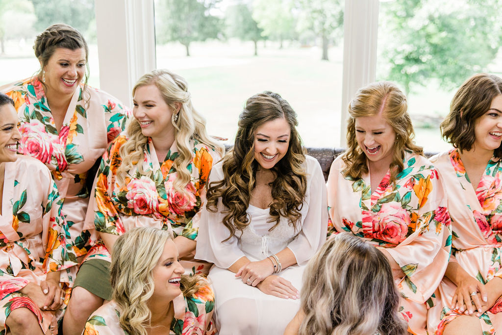 Bridesmaids in their floral robes and the bride in her white robe laughing together. 