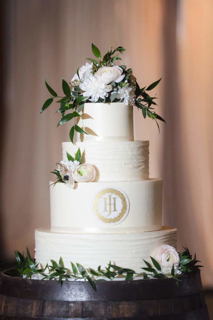 A white, four-tiered cake with white floral, greenery and a custom gold logo