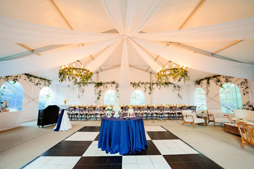 A wide shot of the reception space with a black and white dance floor, white, draping, chandeliers with greenery, soft seating and a long kings table.