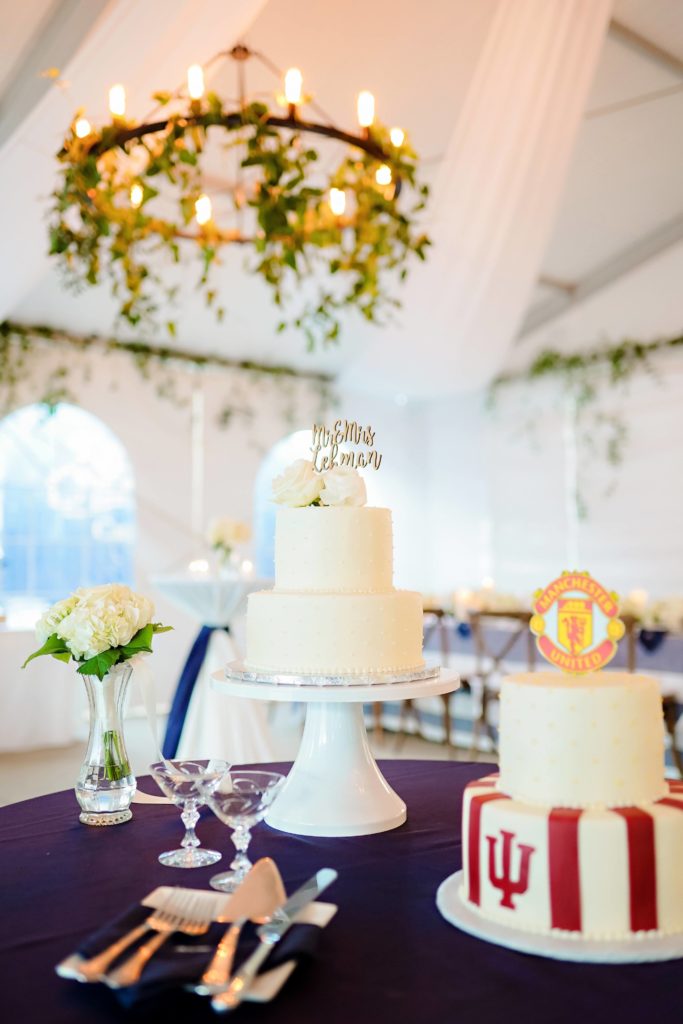 The cake table with navy linen, white floral, and two cakes: a white, two tiered wedding cake and a white and red professional soccer team themed cake. 