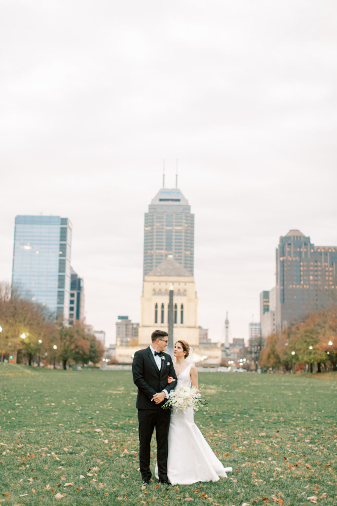 The bride and groom take a photo in the lawn of the Indianapolis central library with downtown Indianapolis in the background