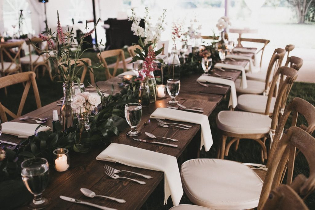 Wooden farm table with wood cross back chairs, greenery running down the center of the table and fresh pink and white flowers and candles throughout the table decor on this private property wedding