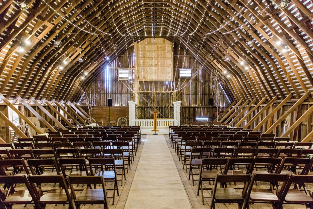 The wedding ceremony set up inside a large barn with exposed wooden beams, located on private property.