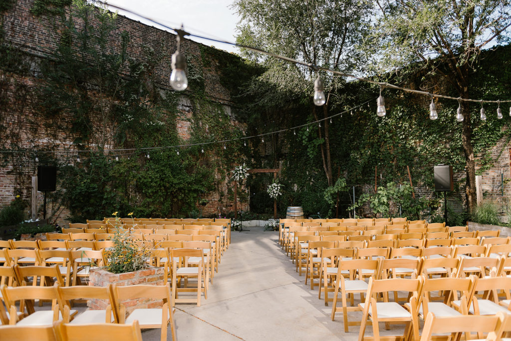 The ceremony had wooden chair, a wooden arh with floral accents, market lighting overhead and a large exposed brick wall background with vines and greenery