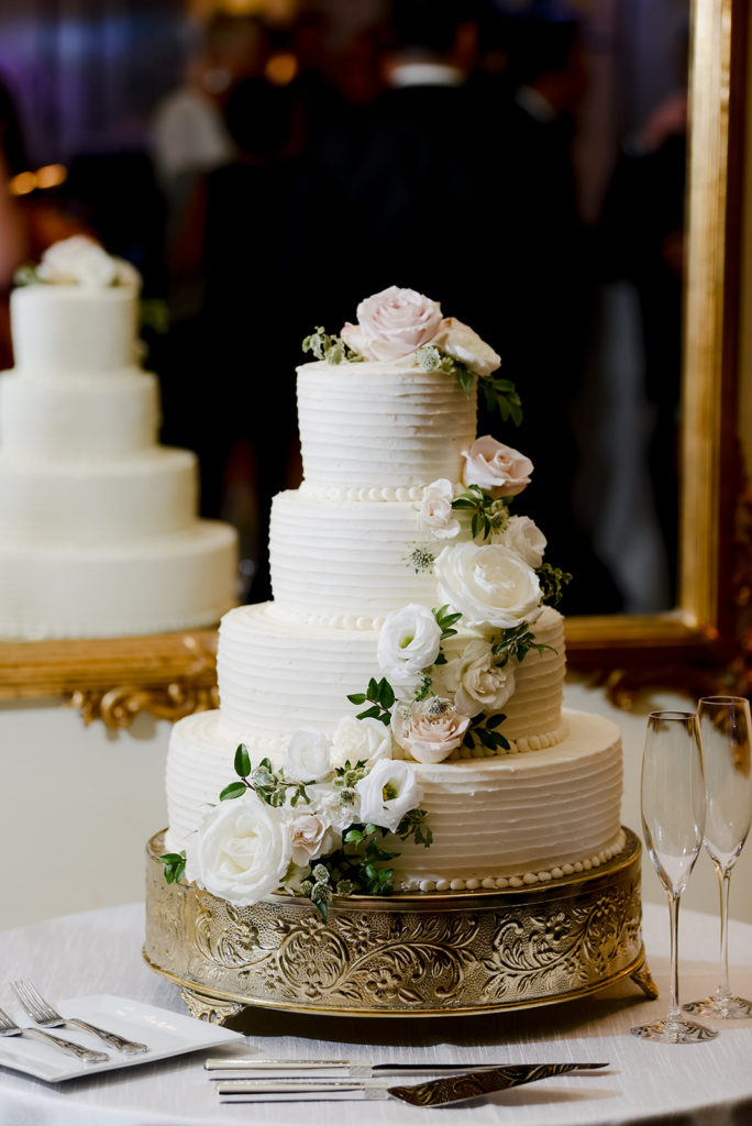 classic white four tiered wedding cake with cascading white and cream flowers placed on a gold cake stand.