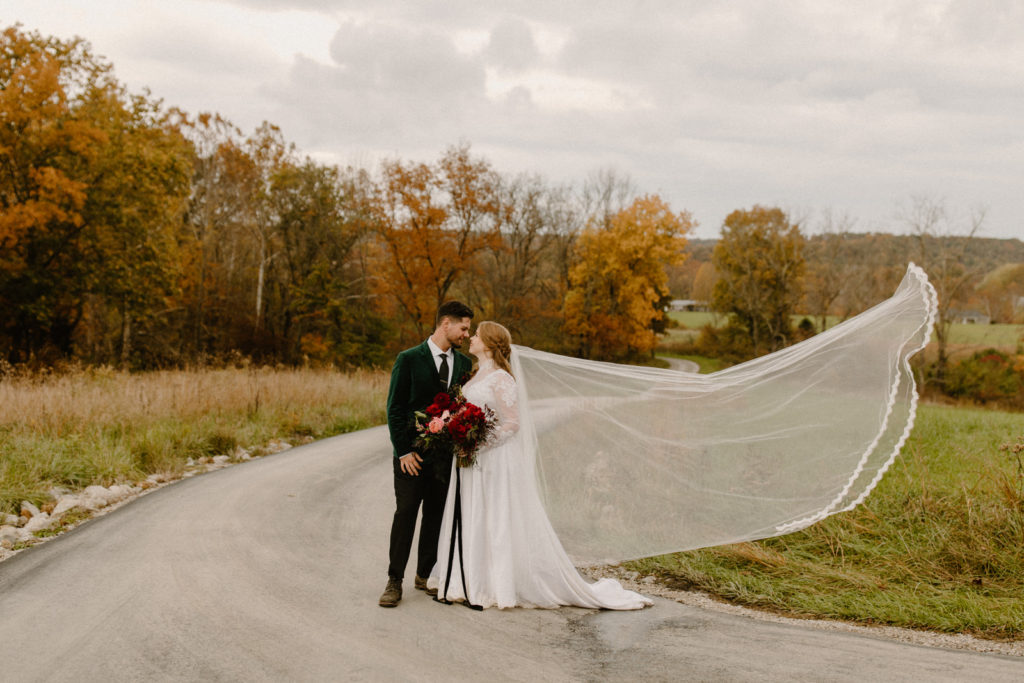 the bride and groom standing on the driveway with orange trees behind them and the brides veil in flows in the wind