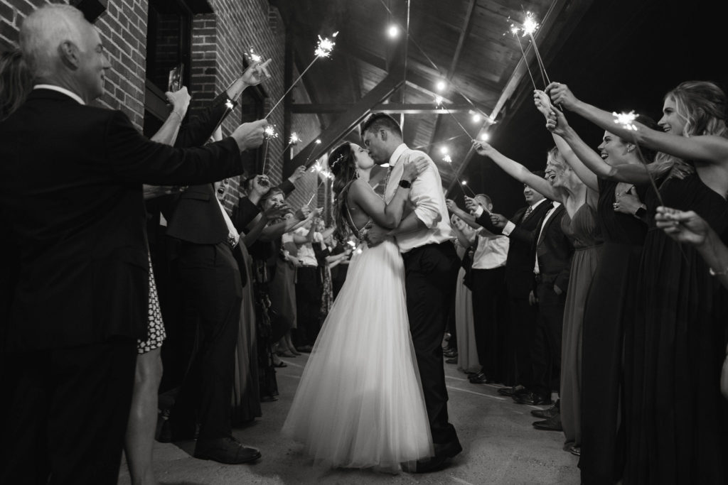 the bride and groom kiss at their sparkler exit with their guests around them in a black and white photo