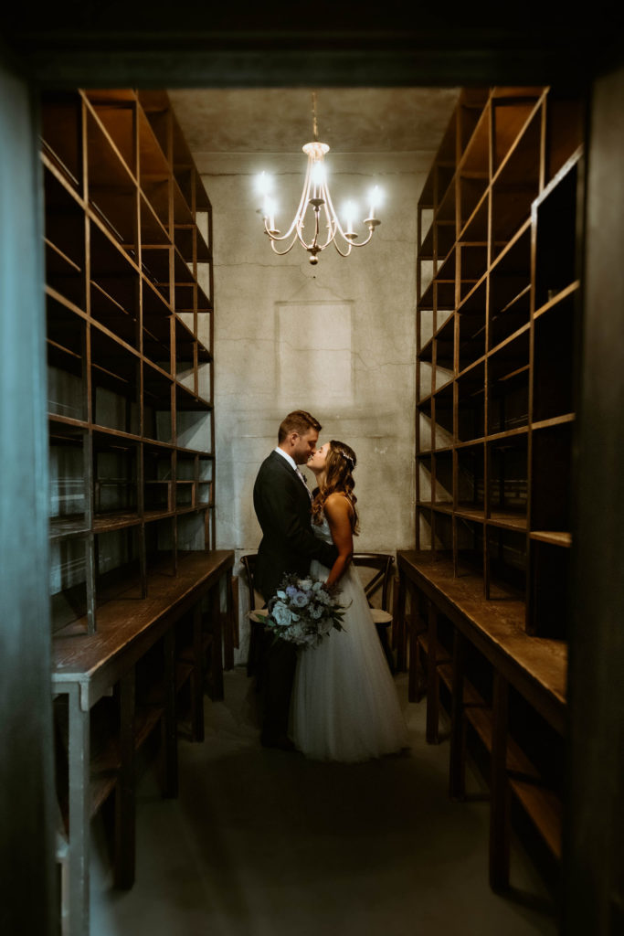 The bride and groom kiss in an empty closet with floor to ceiling wooden shelves and a simple chandelier in the center of the ceiling