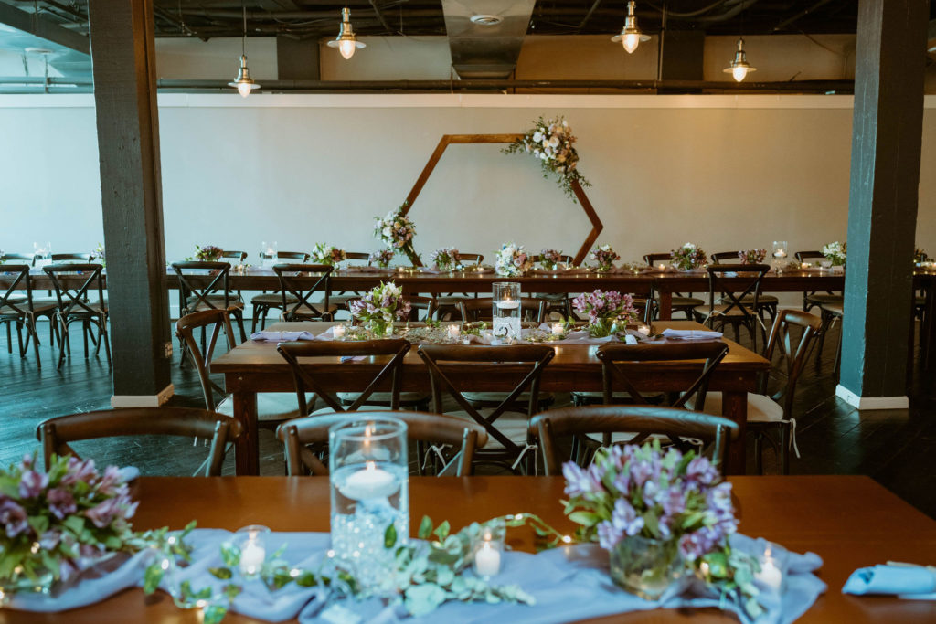 the wedding reception space in the biltwell event center with long wooden tables and chairs with lavender linens, lavender flowers and a hexagon arch with flowers behind the head table 