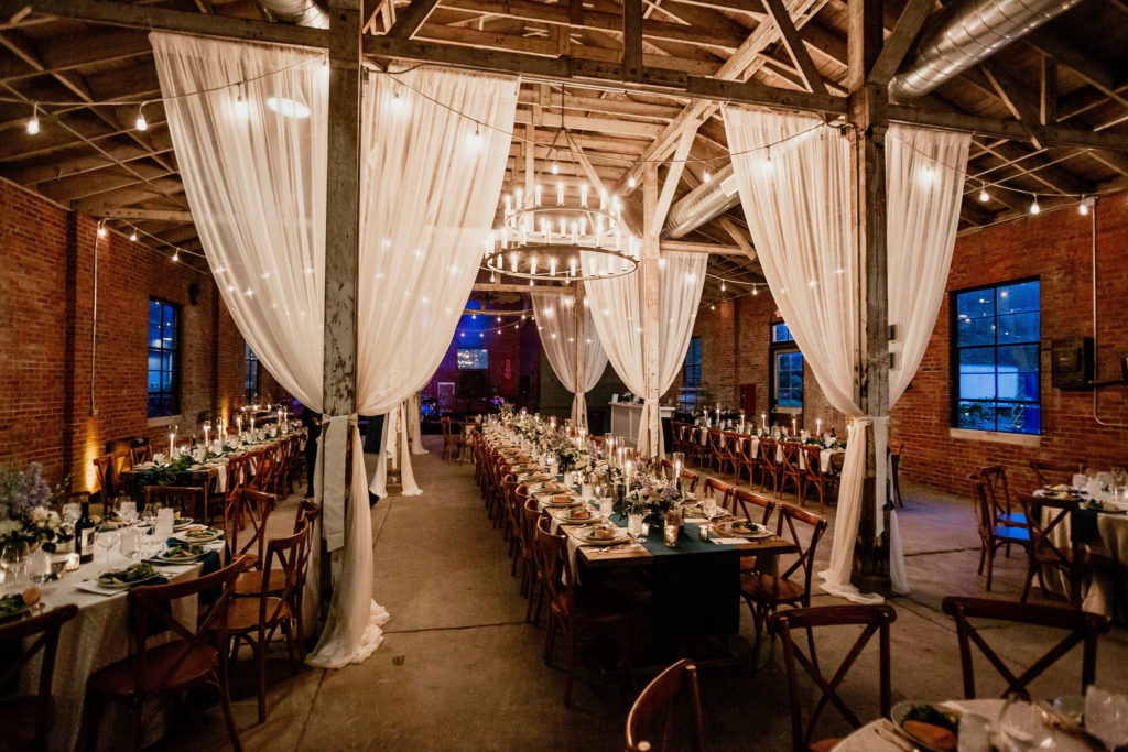 Wedding reception space inside the Castle and Key Distillery venue with brick walls, market lights, white draping and long wooden tables decorated and plated for dinner