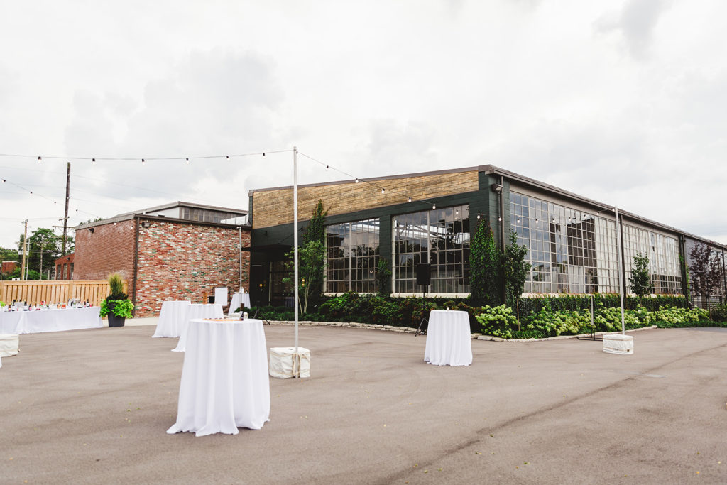 The exterior of the Clerestory wedding venue with white cocktail tables on paved concrete, market lights and greenery lining the black building with large windows