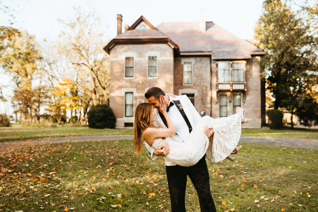 Groom holds the bride in front of historic brick house on the heartland of versailles property: a Lexington KY wedding venue. They are on the front lawn as autumn leaves have fallen around them