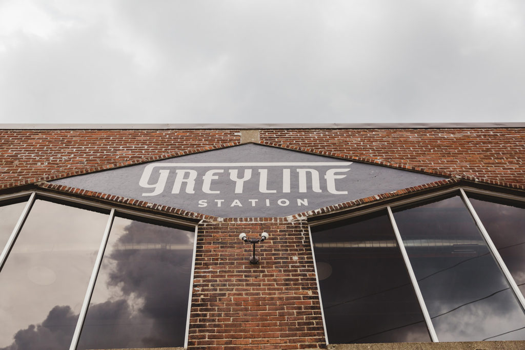 Upward angle of a brick building with two windows and a black sign in the middle of them with white lettering titling the building as the Gremlin Station