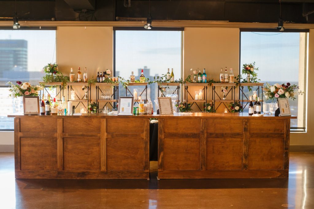 Wooden bar with shelves that hold liquor bottles, glassware and candles with windows overlooking the Lexington city skyline