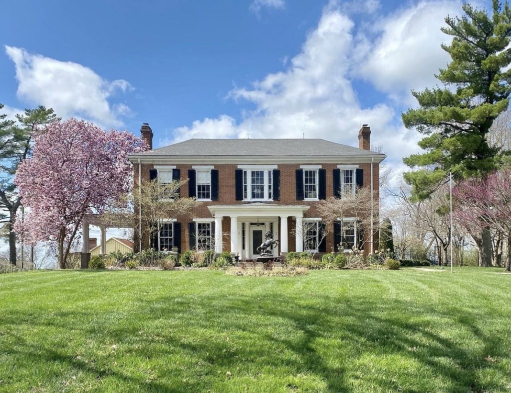 Blue sky and green grass with trees that have pink flowers on both sides of a colonial styled house. The house was red brick with black shutters and white trim with four white columns on each side of the black front door. 