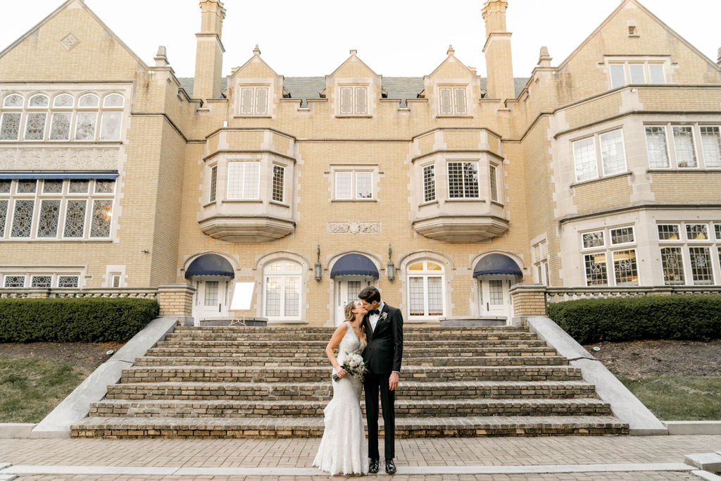 The bride and groom kiss at the base of the exterior steps in front of the grand laurel hall estate on their wedding day