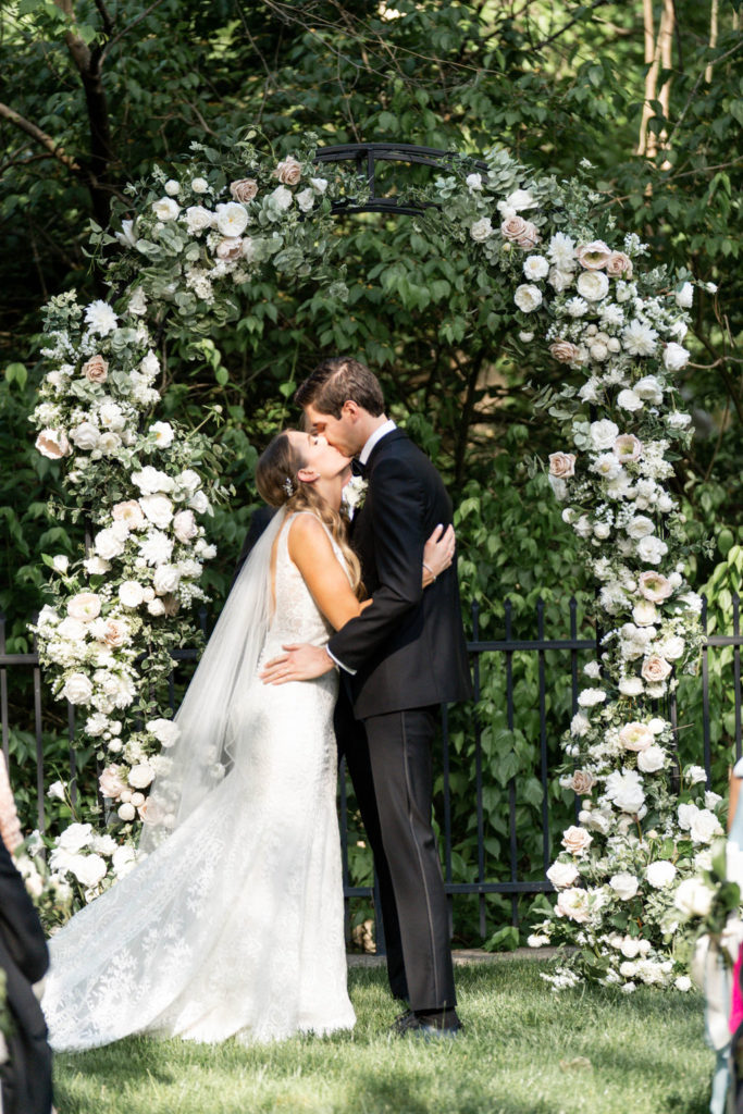The bride and groom kid each other under a white and pale pink flower arch at their outdoor ceremony