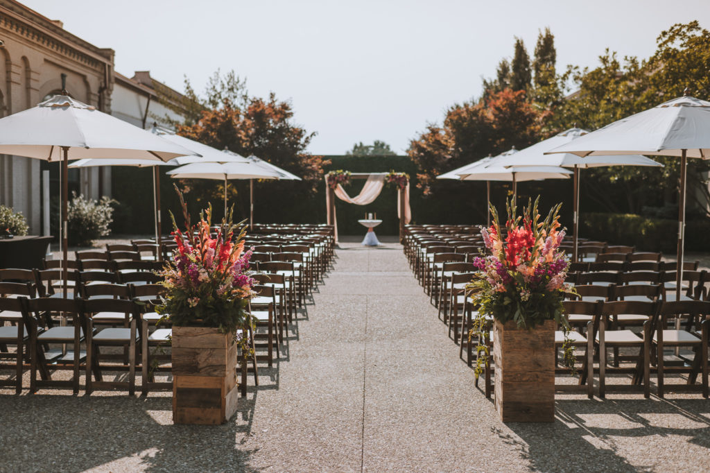 The outdoor ceremony set up in the courtyard of The Refinery in Louisville KY with brown filing chairs, bright flowers on each side of the aisle and white umbrellas for shade.