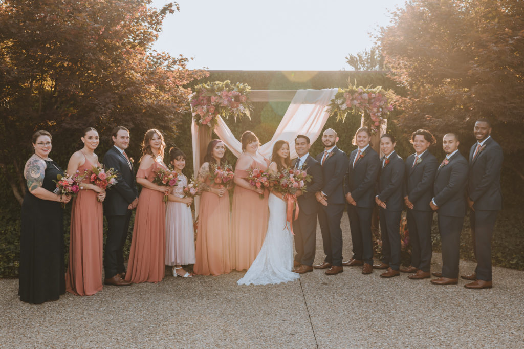 The wedding party smiles together in front of the ceremony arch in the courtyard at The Refinery in Louisville KY