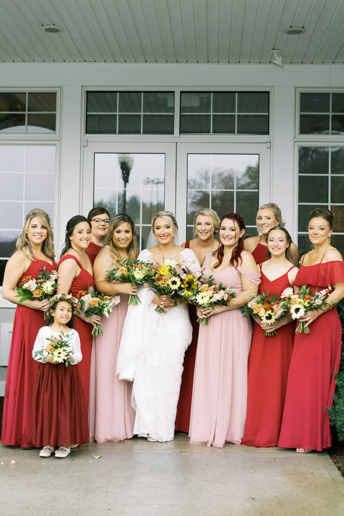 The bride surrounded by her bridesmaids in long red and pink dresses holding fall inspired flower bouquets outside labelle winery
