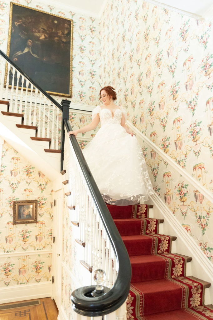 the-bride-walking-down-stairs-lined-with-red-carpet-andcolorful-vintage-wallpaper-on-all-the-walls
