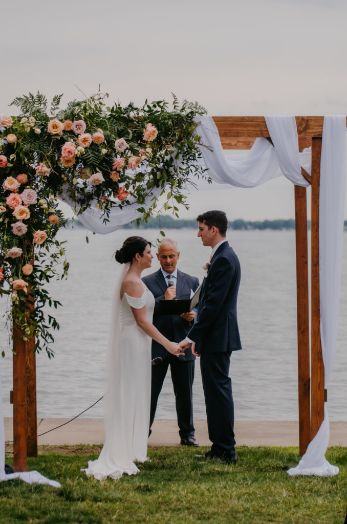 bride and groom hold hands during their lakeside wedding under their ceremony arch 