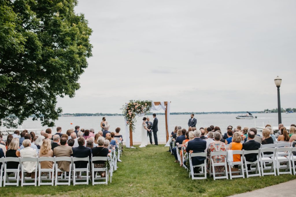 wedding ceremony takes place lakeside on a small grassy lawn, with white chairs and a wood arch with fresh flower arrangements