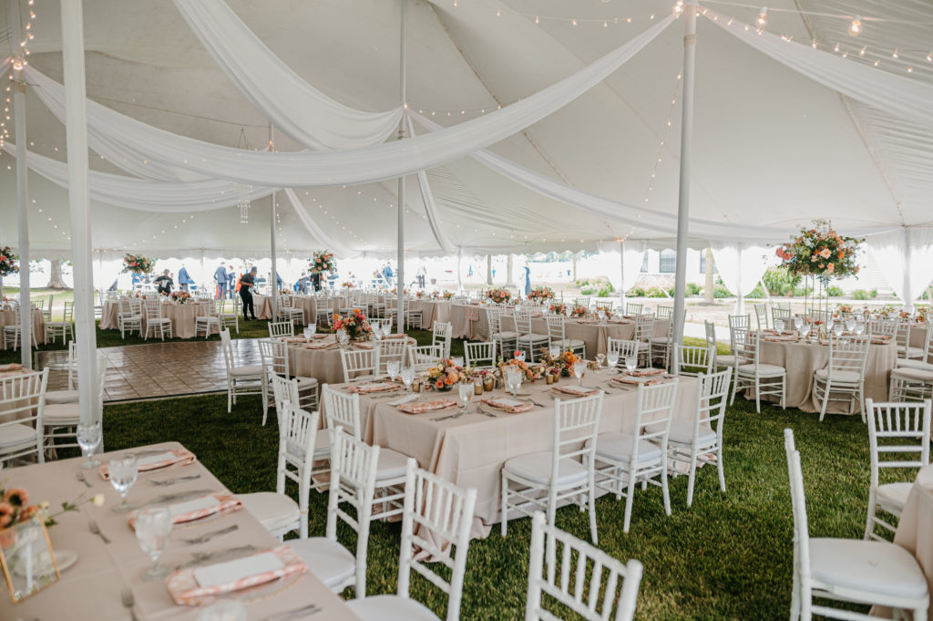 lake wedding reception tent with a neutral tablecloth, white chairs and peach and yellow flowers as table centerpieces 