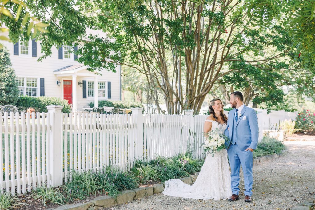 bride and groom look at each other along a white picket fence in front of a White House with a red door and lush greenery