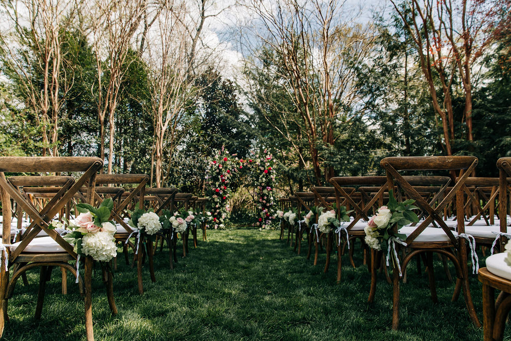 wedding ceremony with wood chairs, white cushions and a big flower arch in a grassy backyard with tall trees