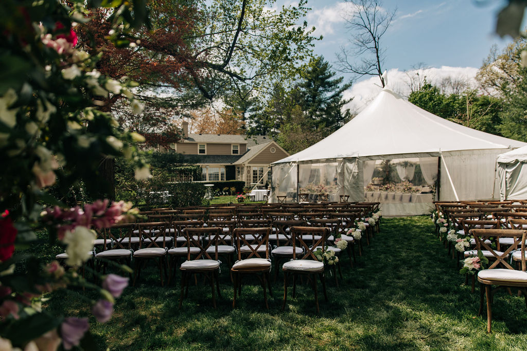 backyard wedding set up with wood ceremony chairs with white cushions, and a large white tent for the reception 