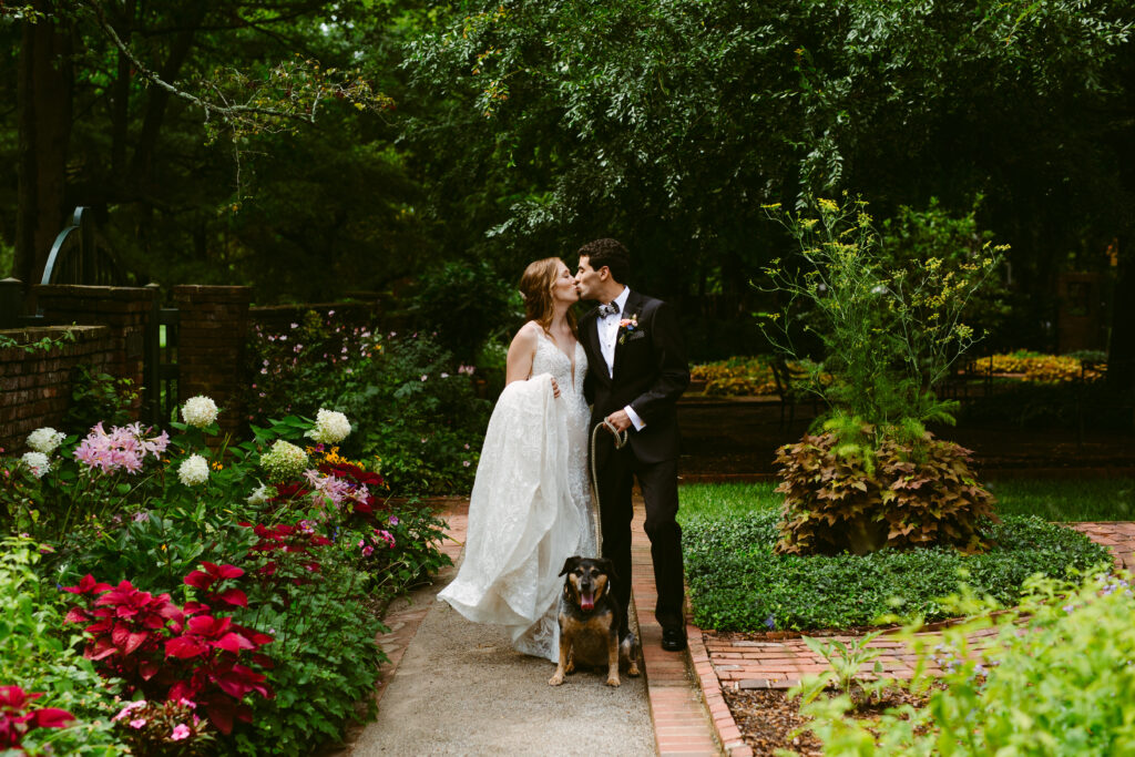 bride and groom kiss and their dog poses for the camera in the gardens of spindletop hall 